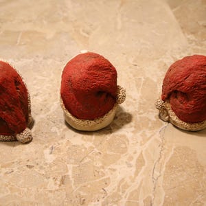 May include: Three small, red Santa hats with a textured appearance and white trim. The hats are arranged on a light brown, marble-like surface, suggesting a festive or holiday theme.