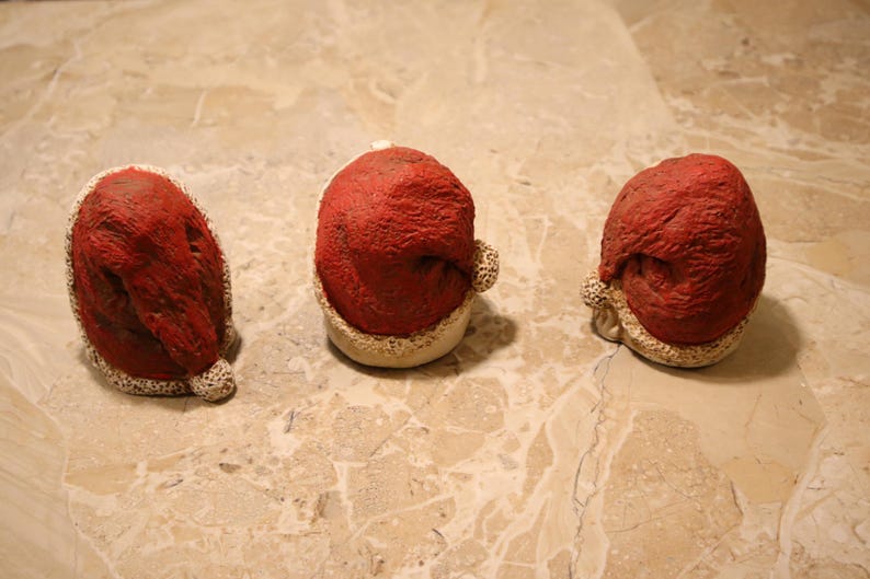 May include: Three small, red Santa hats with a textured appearance and white trim. The hats are arranged on a light brown, marble-like surface, suggesting a festive or holiday theme.