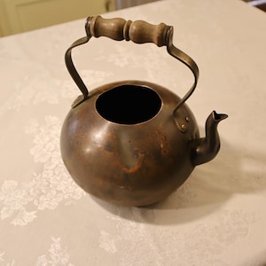 May include: A vintage copper kettle with a round body, a spout, and a wooden handle. The kettle has a dark, aged patina, suggesting it has been used. The handle is made of two wooden pieces. The kettle is sitting on a white tablecloth.