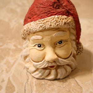 May include: A ceramic Santa Claus head with a red hat and a white beard. The Santa figure has a smiling expression, with rosy cheeks and detailed facial features. The hat has a textured appearance.