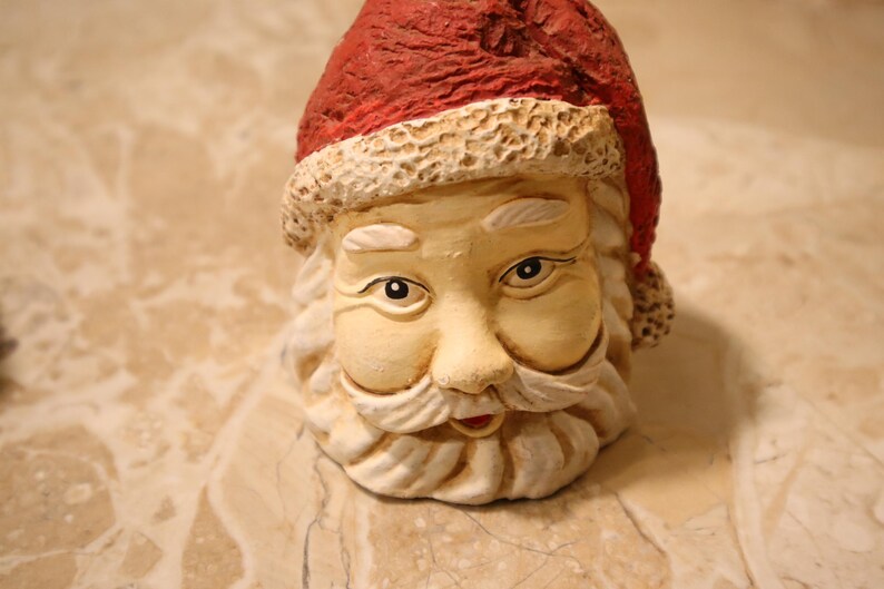 May include: A ceramic Santa Claus head with a red hat and a white beard. The Santa figure has a smiling expression, with rosy cheeks and detailed facial features. The hat has a textured appearance.