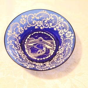 May include: A small, round, cobalt blue glass bowl with a white filigree design around the rim. The center features a white illustration of a bridge and gondola. The bowl is likely decorative.