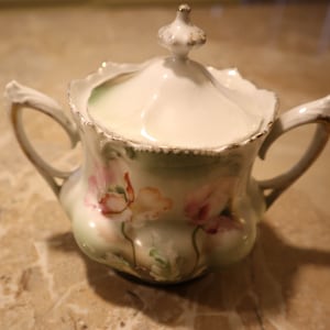 May include: A white porcelain sugar bowl with a lid, featuring a floral design with pink and green accents. The bowl has a gold trim and two handles.