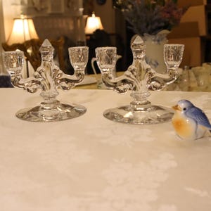 May include: Two clear glass candelabras with three candle holders each. The candelabras have a decorative, ornate design with a round base. A small ceramic bluebird figurine is on the right side of the image.