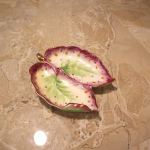 May include: A decorative leaf-shaped dish with a gold stem. The dish features two leaf-shaped compartments with a cream-colored base, green center, and a maroon border with small orange dots. The dish is likely made of ceramic.