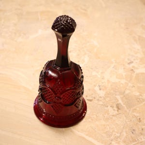 May include: A ruby red glass bell with a textured, rounded top and a long, slender neck. The bell-shaped body features an ornate, embossed design. The bell is approximately 6 inches tall.