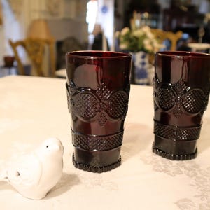 May include: Two deep red glass tumblers featuring a raised, textured geometric pattern around the exterior and a smooth rim. Beside the glasses sits a small, white ceramic bird figurine with a simple, rounded shape and a pointed beak.