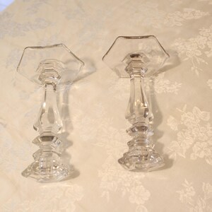 May include: Two clear glass candle holders with hexagonal tops and ornate, faceted stems. The candle holders are displayed on a floral patterned surface, showcasing their elegant design. These decorative pieces are suitable for various home decor styles.