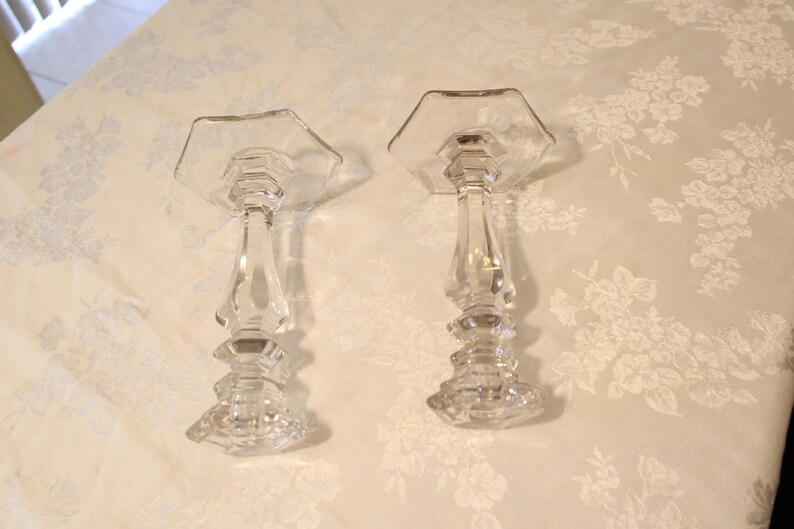 May include: Two clear glass candle holders with hexagonal tops and ornate, faceted stems. The candle holders are displayed on a floral patterned surface, showcasing their elegant design. These decorative pieces are suitable for various home decor styles.