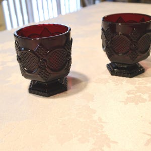 May include: Two ruby red vintage goblets with embossed patterns. The goblets have a square base and a detailed design around the bowl. The interior of the goblets is a deep red colour. The goblets are placed on a floral patterned tablecloth.