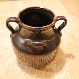 May include: A dark brown ceramic vase with two loop handles and a smaller handle on the front. The vase has a textured, ribbed design on the body and a lighter brown rim.