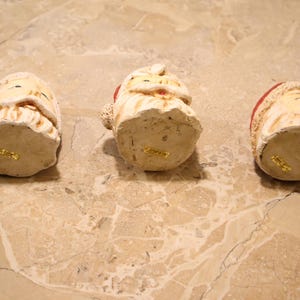 May include: Three ceramic Santa Claus figurines with cream-colored faces, white beards, and red hats. The figurines are arranged on a light brown marble surface. The bottom of each figurine has a gold sticker.