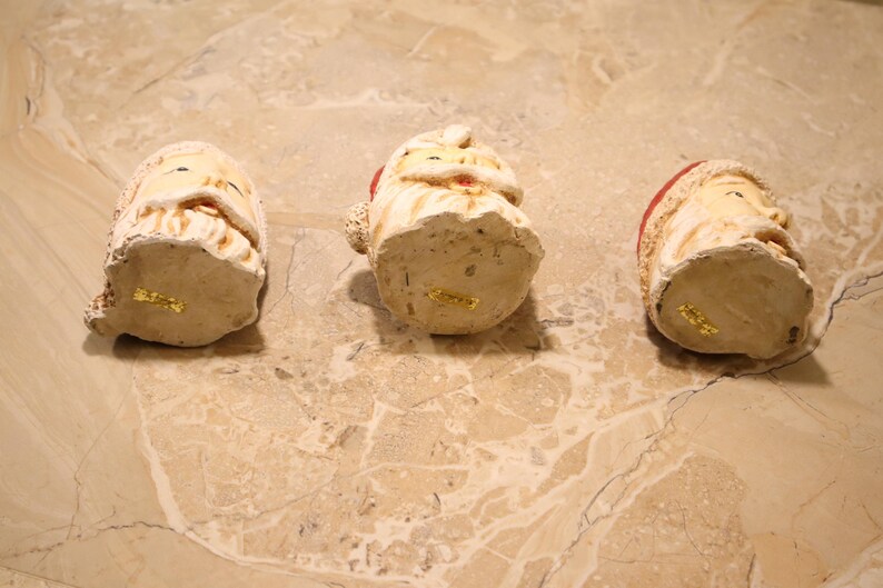 May include: Three ceramic Santa Claus figurines with cream-colored faces, white beards, and red hats. The figurines are arranged on a light brown marble surface. The bottom of each figurine has a gold sticker.