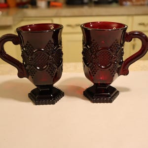 May include: Two ruby red glass mugs with ornate embossed designs and curved handles. The mugs have a pedestal base and are displayed on a white surface. The mugs are likely vintage or antique, and are suitable for serving beverages.