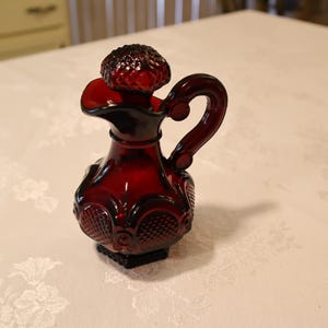 May include: A ruby red glass creamer with a handle and a decorative stopper. The creamer has an ornate, textured design and a small spout. The handle is curved, and the stopper is round with a textured surface. The creamer sits on a square base.