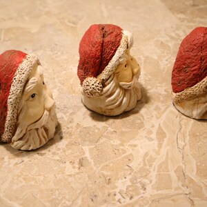 May include: Three Santa Claus head figurines. Each has a red hat with a white trim, a cream-colored face, and a white beard. The figurines are arranged in a row on a light-colored, patterned surface.