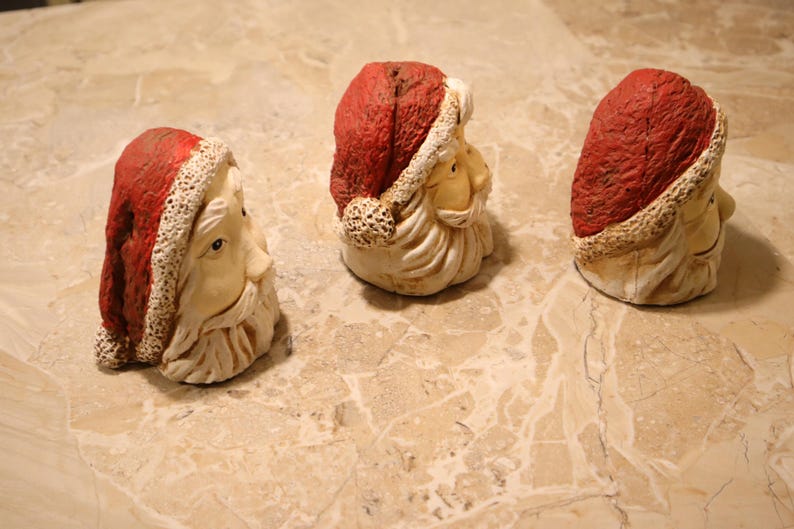 May include: Three Santa Claus head figurines. Each has a red hat with a white trim, a cream-colored face, and a white beard. The figurines are arranged in a row on a light-colored, patterned surface.