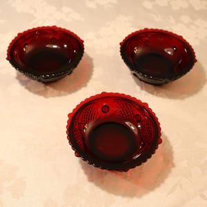 May include: Three small, round, deep red glass bowls featuring a textured pattern along the outer rim. Each bowl has a scalloped edge and a smooth, circular base. The glass is translucent, allowing light to pass through the deep crimson color.