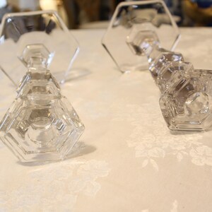 May include: Two clear glass candle holders with a geometric design. The candle holders have a hexagonal base and a faceted stem. The candle holders are on a white floral patterned surface.
