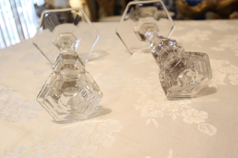May include: Two clear glass candle holders with a geometric design. The candle holders have a hexagonal base and a faceted stem. The candle holders are on a white floral patterned surface.