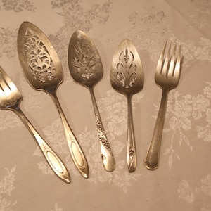 Peut inclure: Cinq ustensiles de service en argent antiques sont disposés sur une surface à motifs floraux. La collection comprend deux fourchettes et trois cuillères de service avec des motifs découpés décoratifs. Les cuillères et les fourchettes ont de longs manches fins.