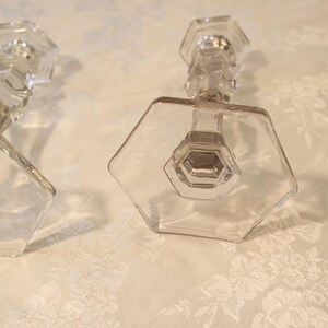 May include: Two clear glass candle holders with hexagonal bases and faceted stems. The candle holders are on a white floral patterned surface. The design is geometric and elegant, suitable for home decor.