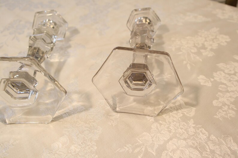 May include: Two clear glass candle holders with hexagonal bases and faceted stems. The candle holders are on a white floral patterned surface. The design is geometric and elegant, suitable for home decor.