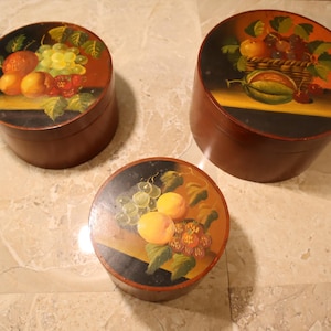 May include: Three round, wooden boxes with lids, each painted with a different still life of fruit. The boxes are brown with black and yellow accents. The fruit includes grapes, peaches, and a watermelon.