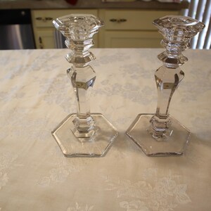 May include: Two clear glass candle holders. Each has a hexagonal base, a faceted stem, and a decorative top. The candle holders are displayed on a white tablecloth with a floral pattern. The background includes a kitchen.
