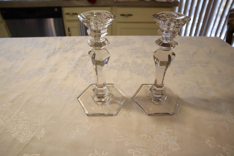 May include: Two clear glass candle holders. Each has a hexagonal base, a faceted stem, and a decorative top. The candle holders are displayed on a white tablecloth with a floral pattern. The background includes a kitchen.