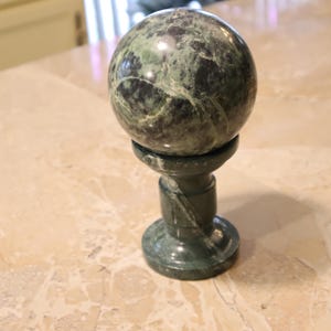 May include: A dark green marble sphere sits atop a matching pedestal. The sphere has a mottled pattern of dark and light green. The pedestal has a round base and a cylindrical stem with a decorative twist.