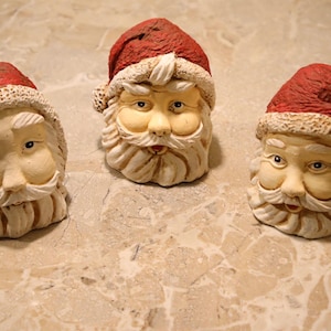 May include: Three Santa Claus head figurines with red hats and white beards. The faces are painted with realistic features, including eyes, noses, and rosy cheeks. The figurines are made of a textured material and are likely decorative items.