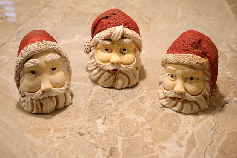 May include: Three Santa Claus head figurines with red hats and white beards. The faces are painted with realistic features, including eyes, noses, and rosy cheeks. The figurines are made of a textured material and are likely decorative items.