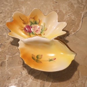 May include: A decorative, two-part ceramic dish in a shell shape. The dish is primarily yellow with an orange gradient and gold trim. It features hand-painted pink and red roses with green leaves. A charming piece for display.