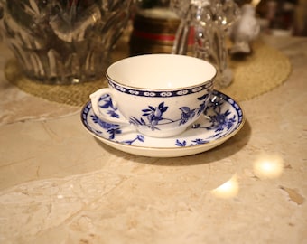 May include: A white teacup and saucer with a blue and gold floral pattern. The teacup has a gold rim and the saucer has a gold band around the edge.