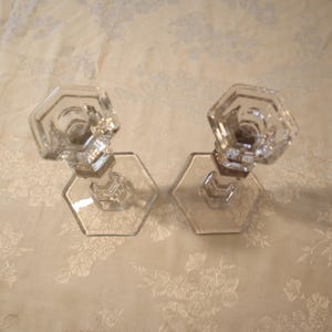 May include: Two clear glass candle holders with hexagonal bases and tops. The candle holders are made of clear glass and are set against a floral patterned background.