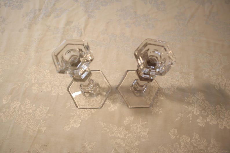 May include: Two clear glass candle holders with hexagonal bases and tops. The candle holders are made of clear glass and are set against a floral patterned background.