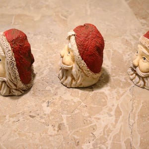 May include: Three Santa Claus head figurines. Each has a red hat with white trim, a white beard, and a smiling face. The figurines are made of a light-colored material and are arranged in a row.
