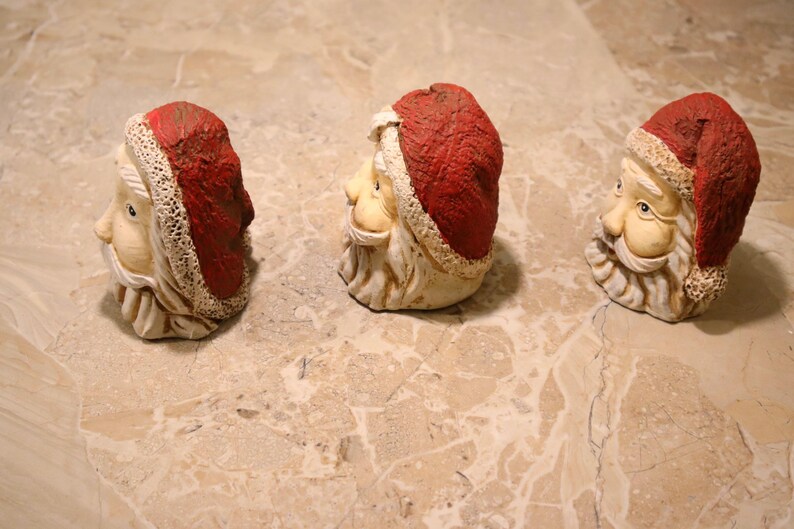 May include: Three Santa Claus head figurines. Each has a red hat with white trim, a white beard, and a smiling face. The figurines are made of a light-colored material and are arranged in a row.
