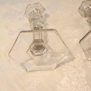 May include: Two clear glass candle holders with hexagonal bases and stems. The candle holders have a geometric design and are placed on a white floral patterned surface. The glass appears to be vintage.