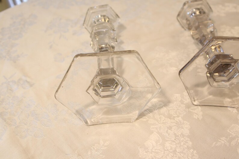 May include: Two clear glass candle holders with hexagonal bases and stems. The candle holders have a geometric design and are placed on a white floral patterned surface. The glass appears to be vintage.