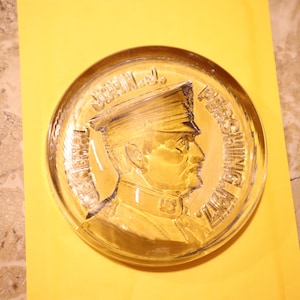 May include: A clear glass paperweight with a raised image of a man in a military uniform. The text "U.S. NAVY PERSHING" is inscribed around the edge of the paperweight.