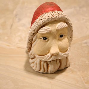 May include: A ceramic Santa Claus head figurine. The Santa head has a red hat with a white trim, a long white beard, and a neutral skin tone. The figurine is a festive decoration.
