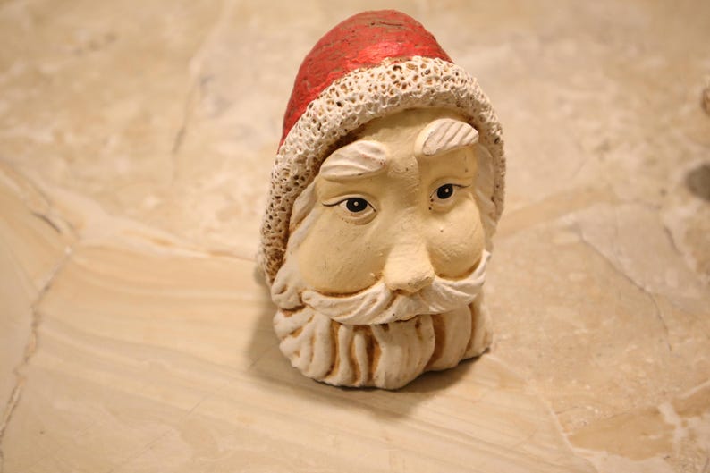 May include: A ceramic Santa Claus head figurine. The Santa head has a red hat with a white trim, a long white beard, and a neutral skin tone. The figurine is a festive decoration.