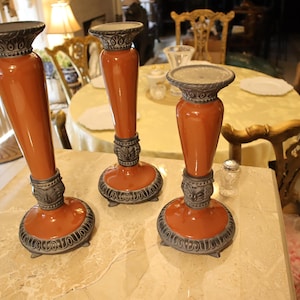 May include: Three decorative candle holders. Each holder has a burnt orange body with a silver-colored base and top. The holders are arranged on a marble-like surface, with ornate furniture in the background.
