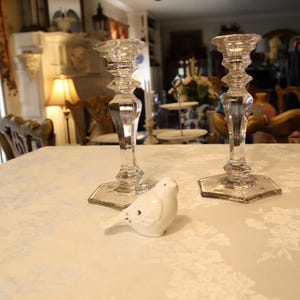 May include: Two clear glass candle holders with a hexagonal base and a small white ceramic bird on a white tablecloth. The candle holders have a detailed, faceted design. The table is covered with a white floral pattern.