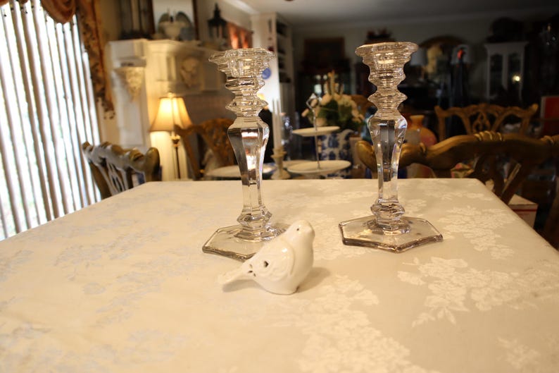 May include: Two clear glass candle holders with a hexagonal base and a small white ceramic bird on a white tablecloth. The candle holders have a detailed, faceted design. The table is covered with a white floral pattern.