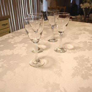 May include: A set of three clear glass goblets with smooth bowls and twisted stems. The glasses are arranged on a white tablecloth featuring a subtle, tone-on-tone floral damask pattern. Each goblet has a circular base and a slender, elegant profile.