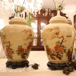 May include: Pair of cream-colored ceramic table lamps. Each lamp features a floral and bird design in shades of orange, brown, and green. The lamps have a gold base and a black wooden base. The lamps are connected to a power source.