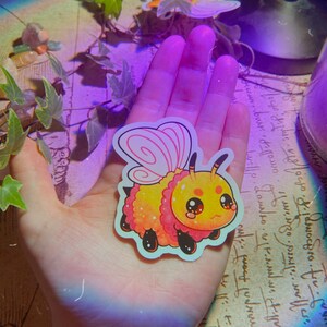 Op de afbeelding: Een gele en roze pluche bij met een roze vlinder vleugel sticker. De bij heeft een schattig cartoon gezicht met grote ogen en een kleine glimlach.